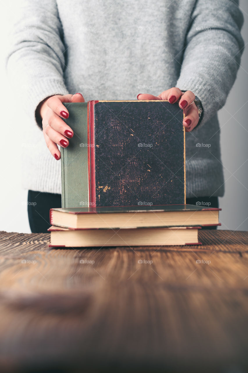 Young woman holding a few old books on wooden table in antique bookstore. Woman wearing grey sweater and jeans. Vertical photo