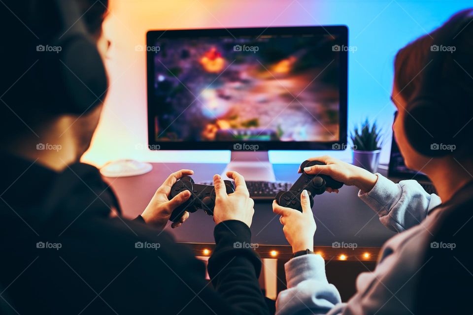 Friends playing video game at home. Gamers holding gamepads sitting at front of screen. Streamers girl and boy playing online in dark room lit by neon lights. Competition and having fun