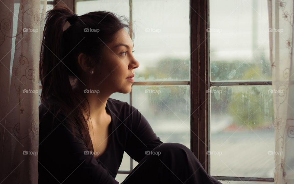 Young woman sitting on her window looking outside of the house with hopeful eyes 