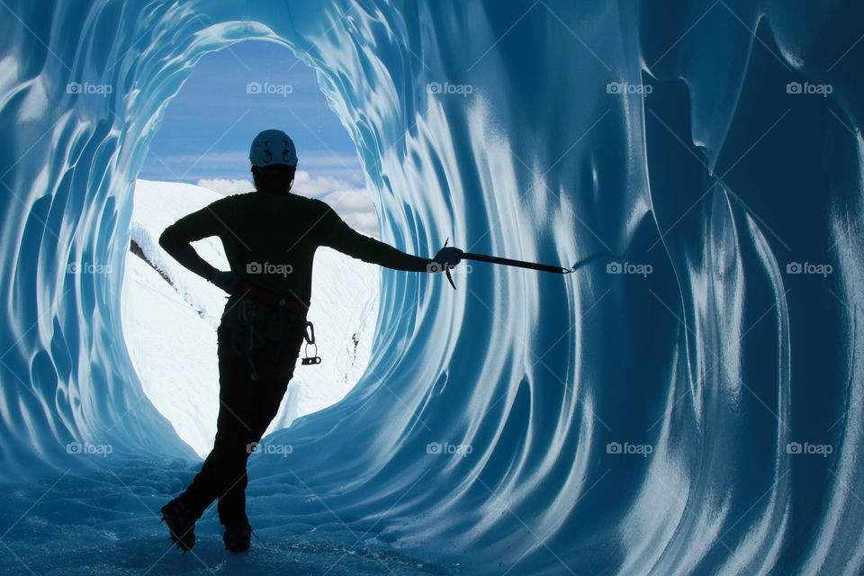 Man standing leaning against ice axe on the wall of an ice cave in the Matanuska Glacier in Alaska. He has a helmet, harness, and belay device for ice climbing. Legs crossed and hand on hip.