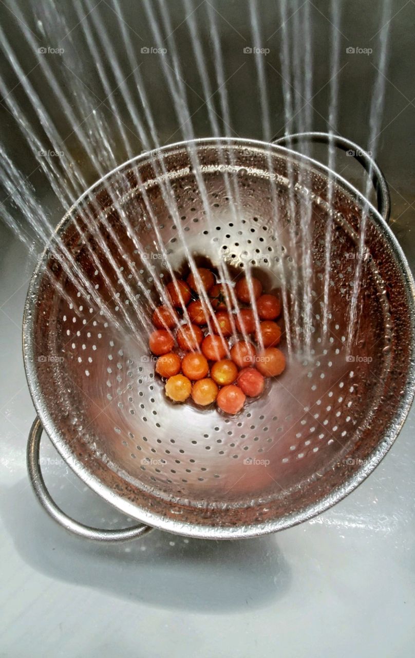 Kitchen sink faucet spraying water into silver metal colander that is holding ripe cherry tomatoes.