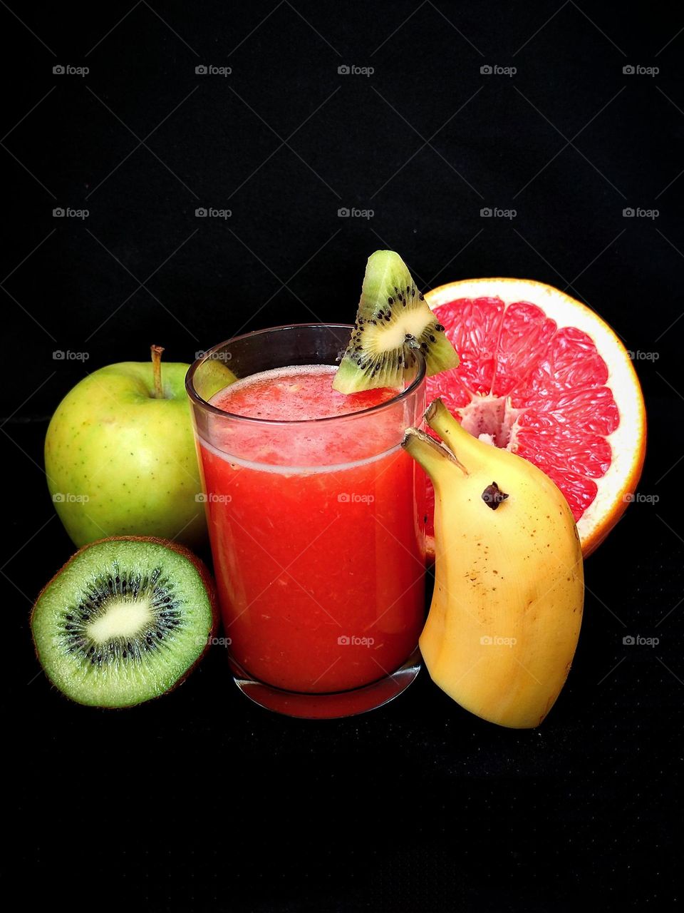 Food. On a black background a glass with red juice.On the edge of the glass is a triangle of green kiwi. Juice ingredients lie around the glass: half a green kiwi, a green apple, half a red grapefruit, half a yellow dolphin-shaped banana