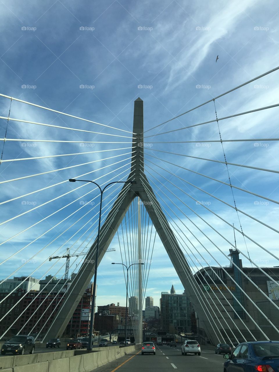 Far away pic of Leonard Zakim Bridge showing more of its detail. City of Boston MA🇺🇸all around. Highway on both sides, blue sky, white Bridge.