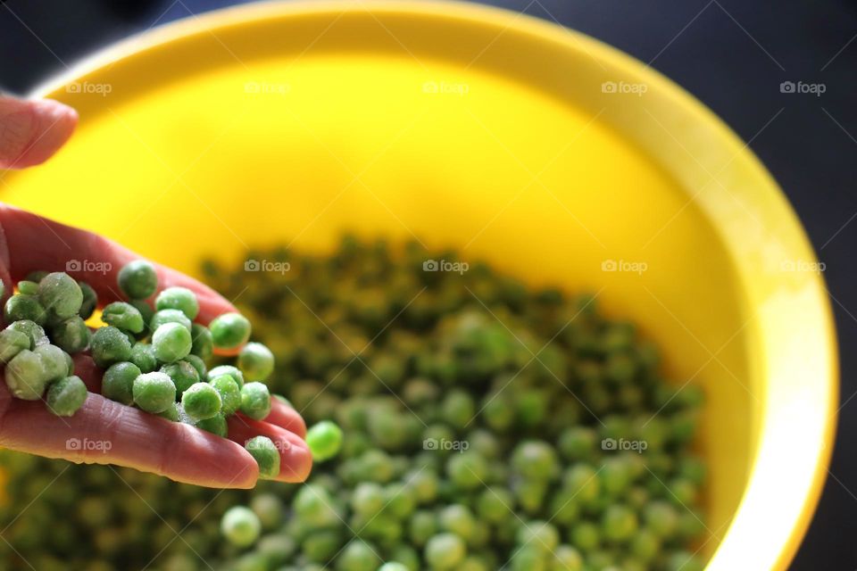 A hand holds green peas over a yellow bowl of peas