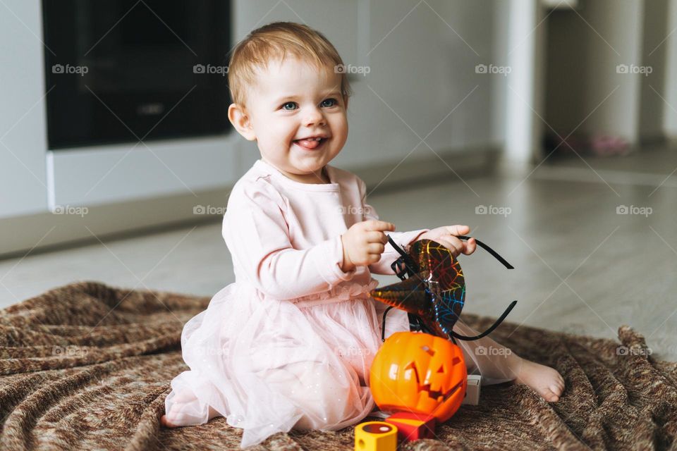 Cute little baby girl in pink dress with the witch's hat and pumpkin lantern sitting on floor in kitchen at the home, Halloween time