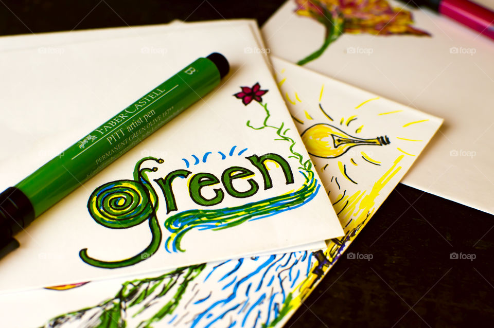 Faber-Castell artist pen colors olive green on desk with word “green” written in nature design on card stock next to light bulb a creative images conceptual ideas, earth day, green planet environmental words and images photography background 