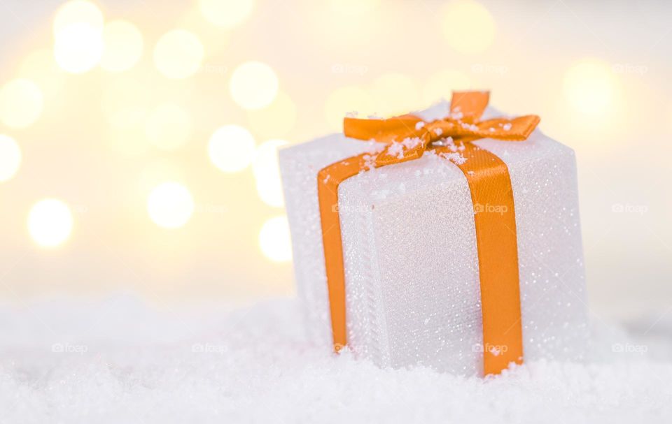 One small gift box with an orange ribbon lies on the right in the snow on a blurred background with bokeh, close-up side view. Holiday banner concept, holiday gifts.