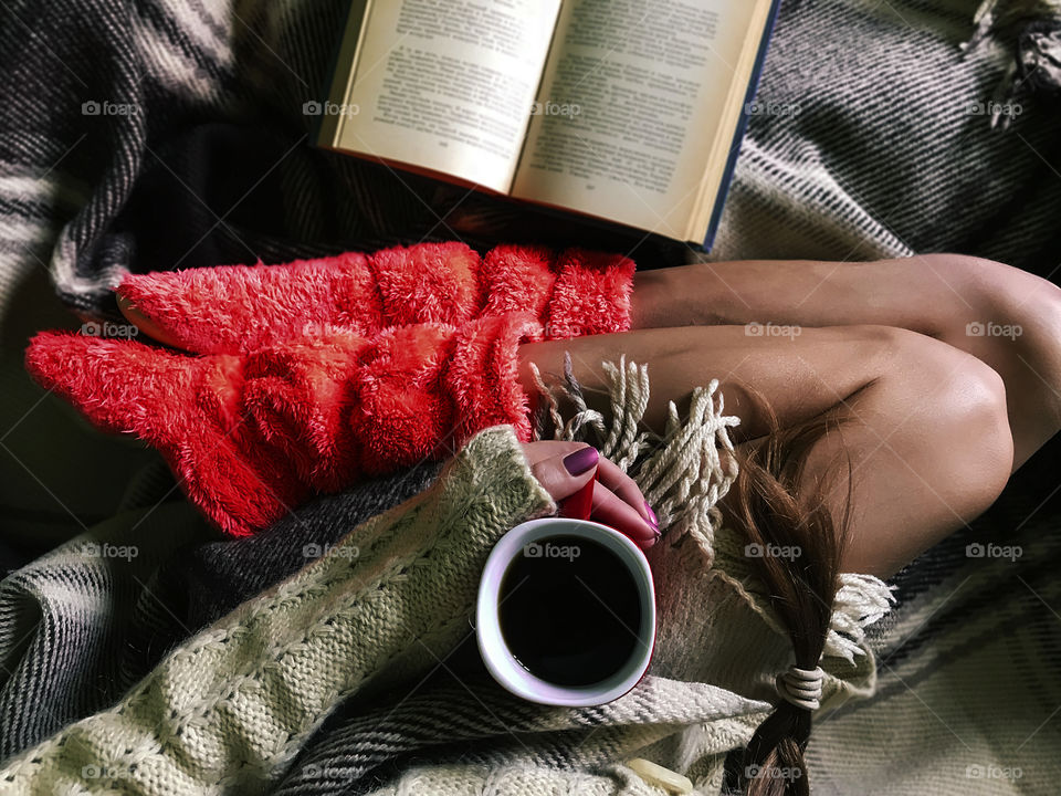 Young woman wearing sweater and cozy pink home boots enjoying coffee and reading a book in cozy bed 