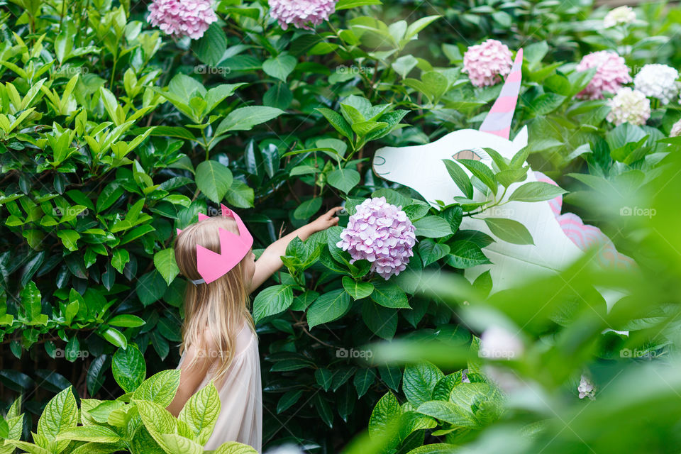 Happy little Caucasian girl with blonde hair and crown standing near unicorn in blooming garden at summer day