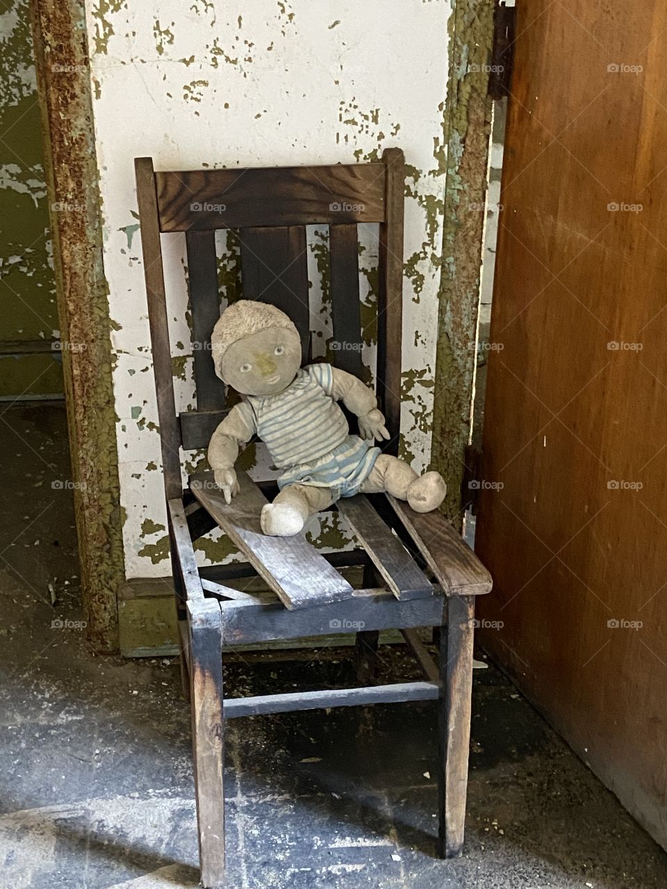 An old creepy rag doll sitting on a broken wooden chair in front of a decaying wall