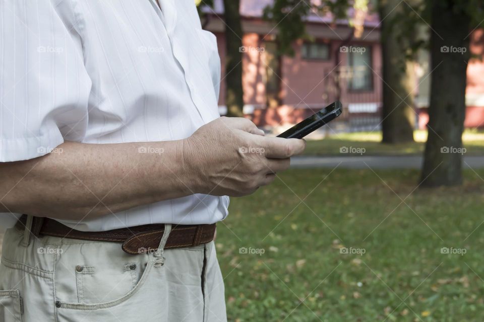 An elderly man walks alone in the park in the summer. A modern pensioner, businessman in a white shirt and trousers takes pictures with a camera in a mobile phone.