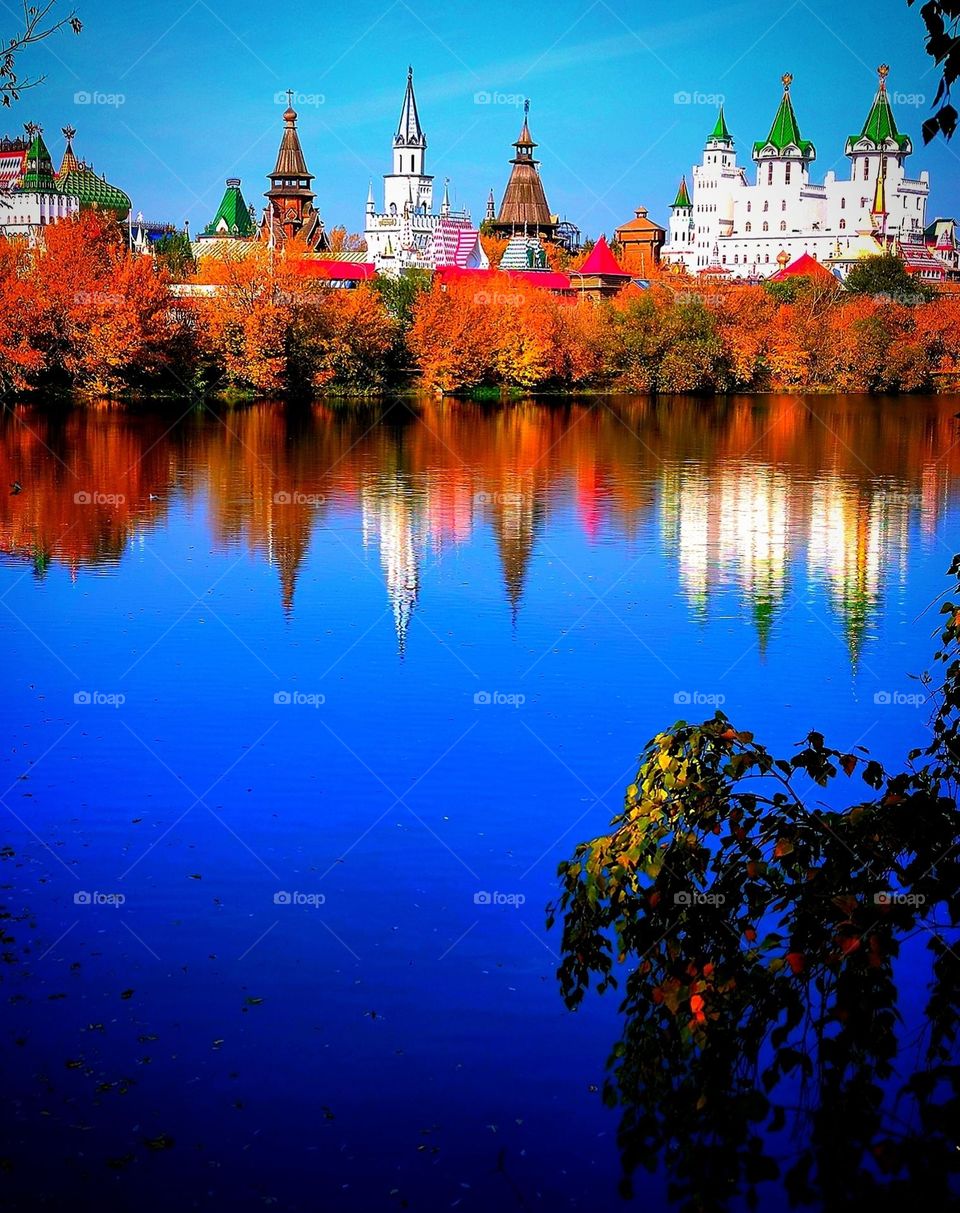 Autumn landscape. On the bank of the river, among the autumn trees, the domes of the towers are visible. Trees and domes of towers are reflected in the dark blue water. In the foreground is a birch branch with green and yellow leaves.