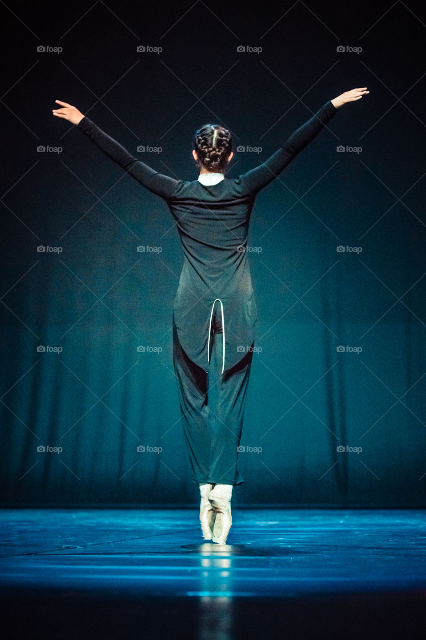 Young Woman Ballerina Dancer Raises Hands Up On Stage In Dark Background
