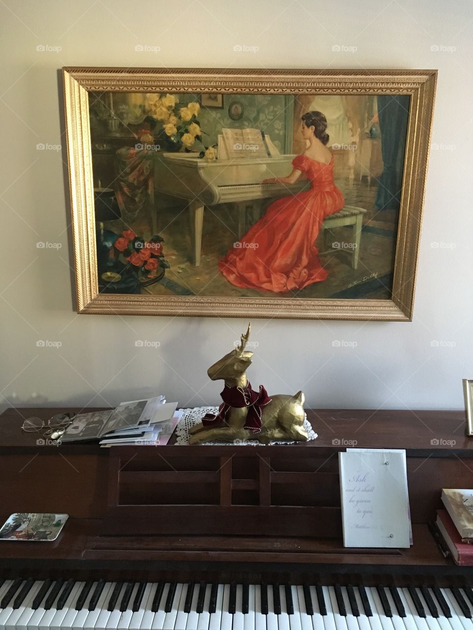 An old grand piano covered in letters and papers with a golden reindeer decoration and a beautiful painting of a woman playing the piano surrounded by flowers