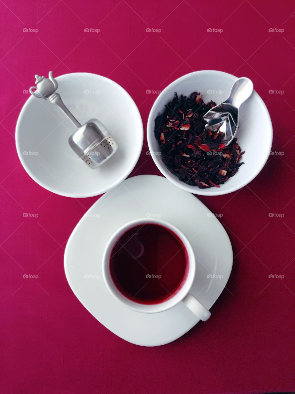 Tea Time - flat lay, triangular arrangement of stainless steel teapot steeper in white bowl, dried hibiscus flowers in white bowl, hibiscus tea in white cup on magenta fabric background