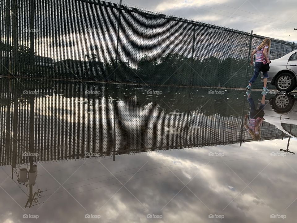 Urban fence and cloudy sky reflected in puddle as girl walks to car