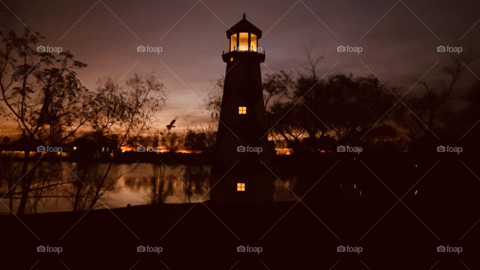 Lighthouse is standing by. Sky is multicolored with layers of high clouds not letting good light through. Reflections of shore line can be seen mirrored on Lake Waters. 
