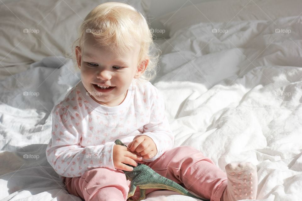 Happy baby girl with toy laughing on white bed in the lights of a sun