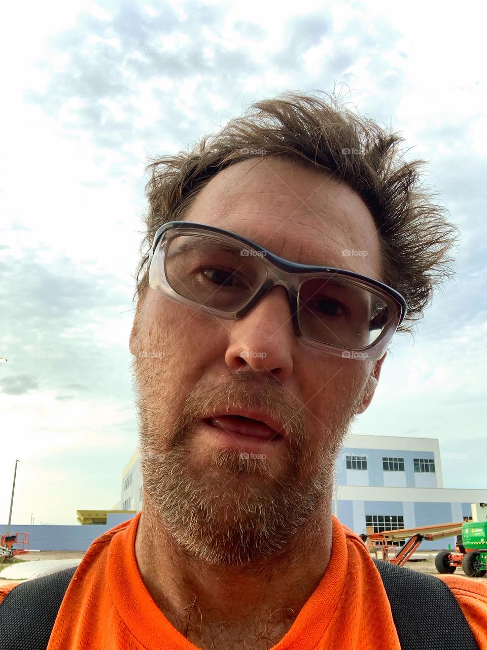 Selfie hard worker under heat in carpentry building scaffold showing heat exhaustion in a funny way with humor with safety glasses and machinery in front of large building.