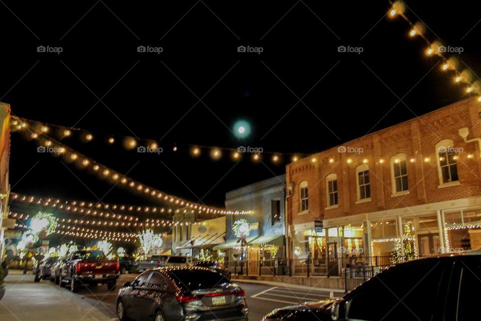 Antiquated downtown buildings are decorated with string lights down the street. The moon can be seen just above the lights, helping bathe the city in light