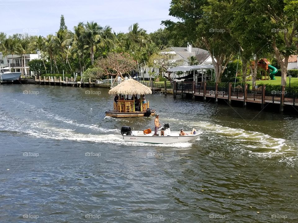 Boats on river, luxury home in background on ocean rivers shore. Two different styles of boats going down river way or canal. People on bar stools drinking. It's a hot sunny &  humid day. Tropical scene.