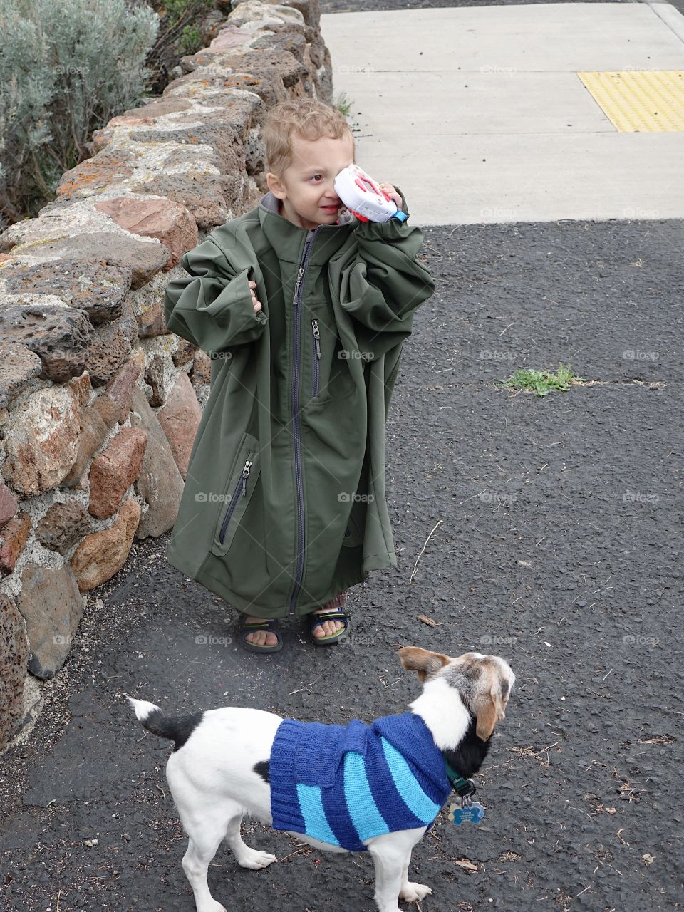 A cute little boy draped in an adult jacket with his sweater clad small dog plays at taking a picture while holding his toy camera backwards on a cold spring day in Central Oregon. 