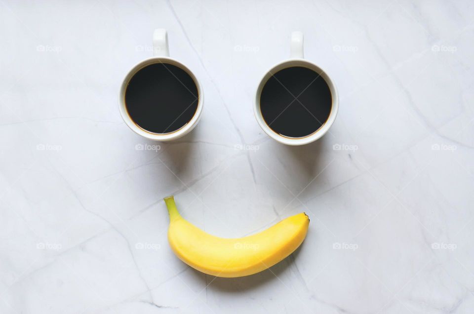 Top view of yellow smiling banana and two cups of black coffee close up. White background.  Food and drinks