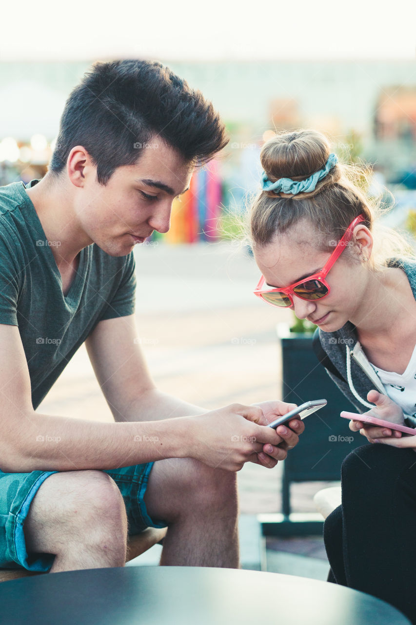 Couple of friends, teenage girl and boy, having fun using smartphones sitting in center of town, spending time together