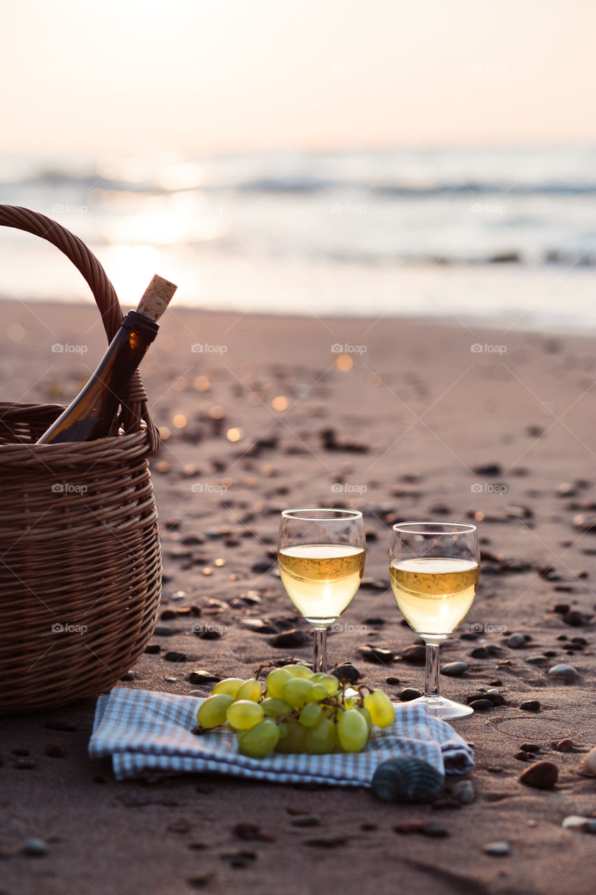 Two wine glasses with white wine standing on sand, on beach, beside grapes and wicker basket with bottle of wine. Sea waves in the background