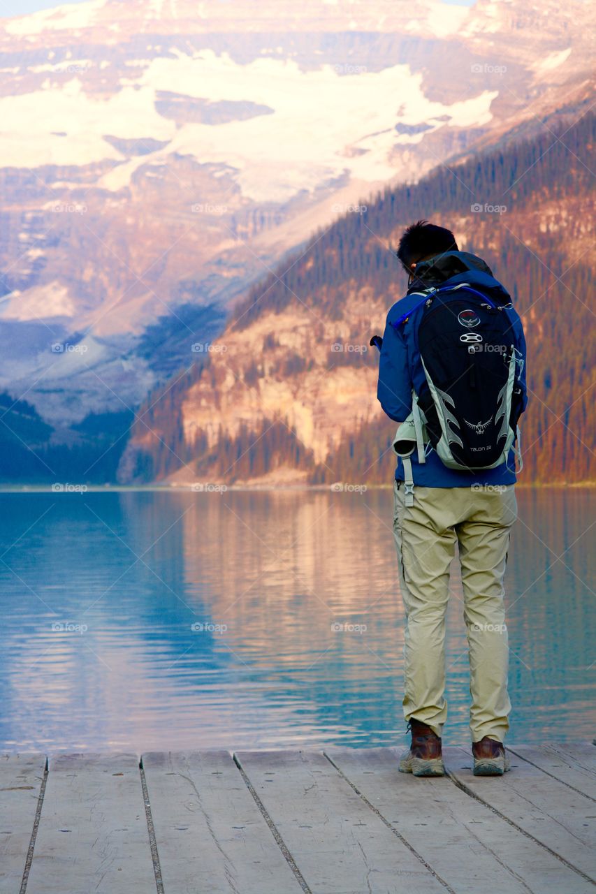 Young man in backpack photographing beautiful glacial lake and snow capped mountains of stunning Lake Louise near Banff Alberta in Canada's Rocky Mountains 