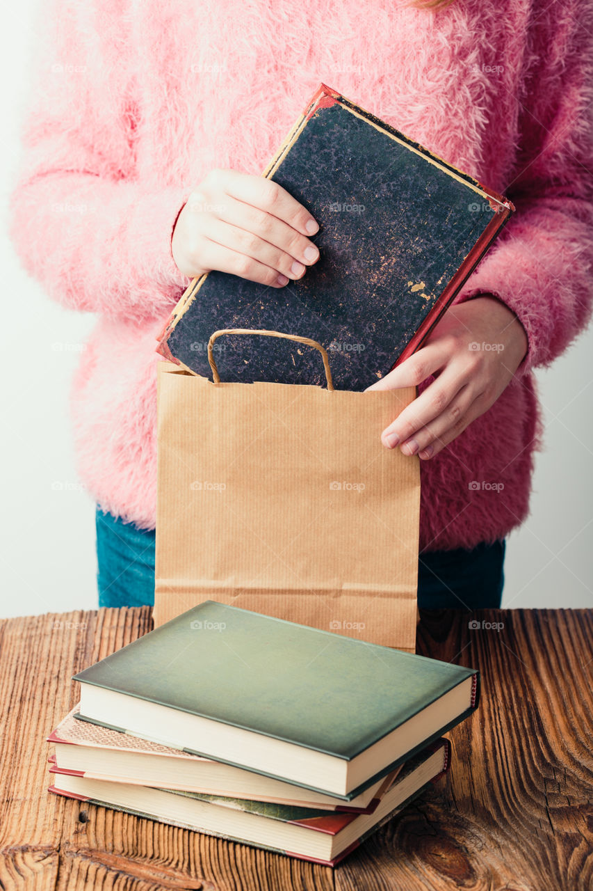 Young girl turning the pages of book in bookstore. Putting the books into paper bag. A few books on a wooden table. Teenager girl wearing pink sweater and blue jeans. Vertical photo