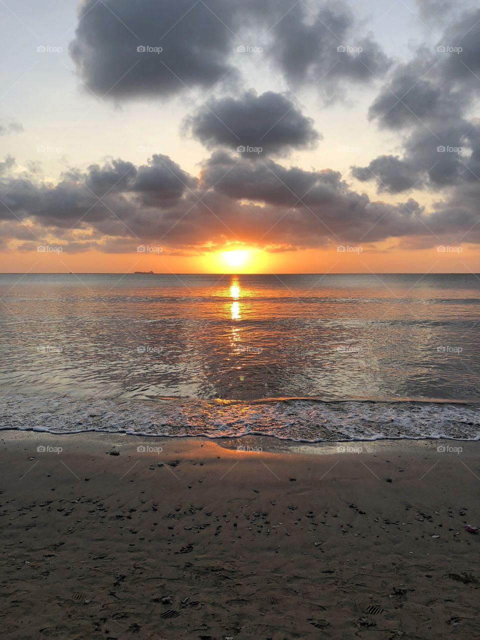 Cloudy sunset on Jimbaran beach. Orange glow, calm water. Distant ship on horizon. Bali, Indonesia. 2018.