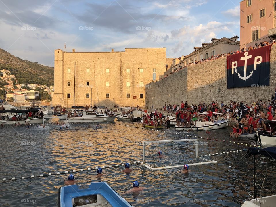 This is a picture of a water polo game in the old Harbour In Dubrovnik Croatia featuring the water and fans in the distance