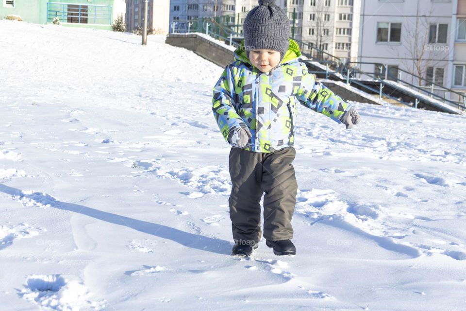 A child in winter in winter jackets, pants, a hat and boots on white snow on the street and in the park in nature plays winter fun and sleds.