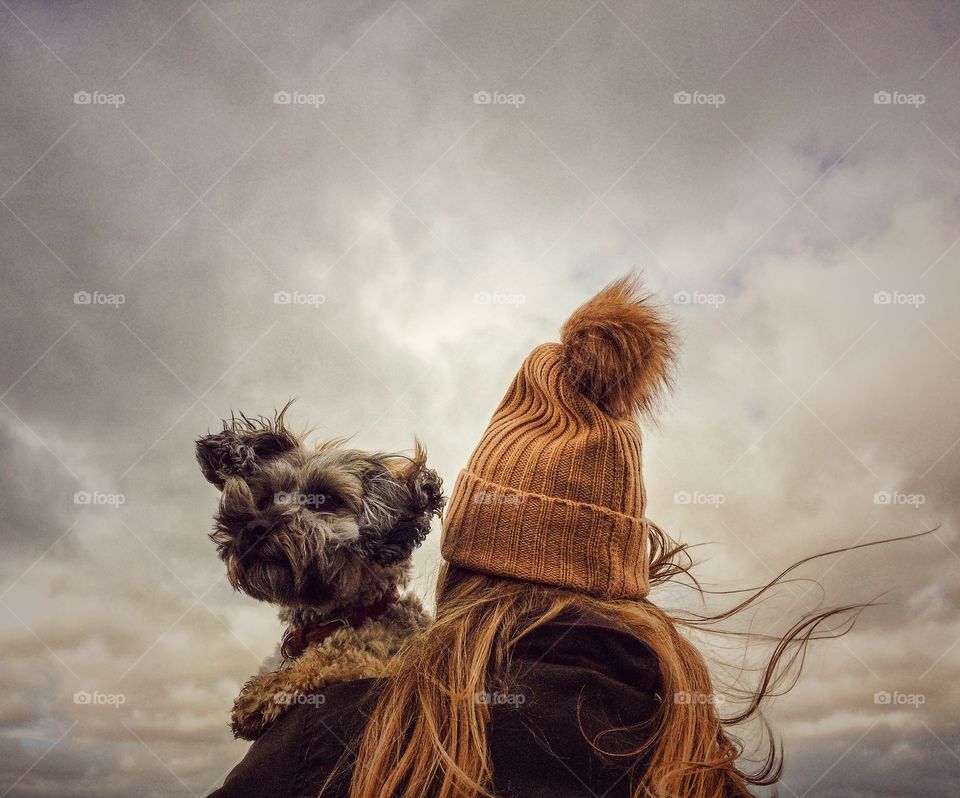 A rear view of fashionable young girl wearing a woolly bobble hat and holding her scruffy pet dog on her shoulder on a windy day in autumn 