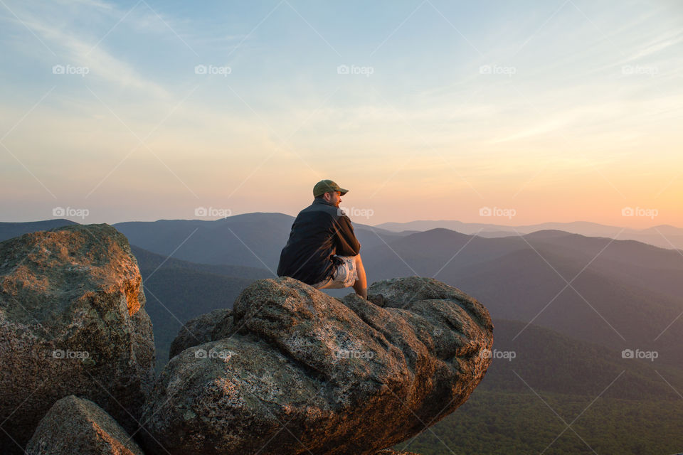A man kneels on the top of Old Rag Mountain, VA, surveying the Blue Ridge Mountains and Shenandoah National Park at dawn.