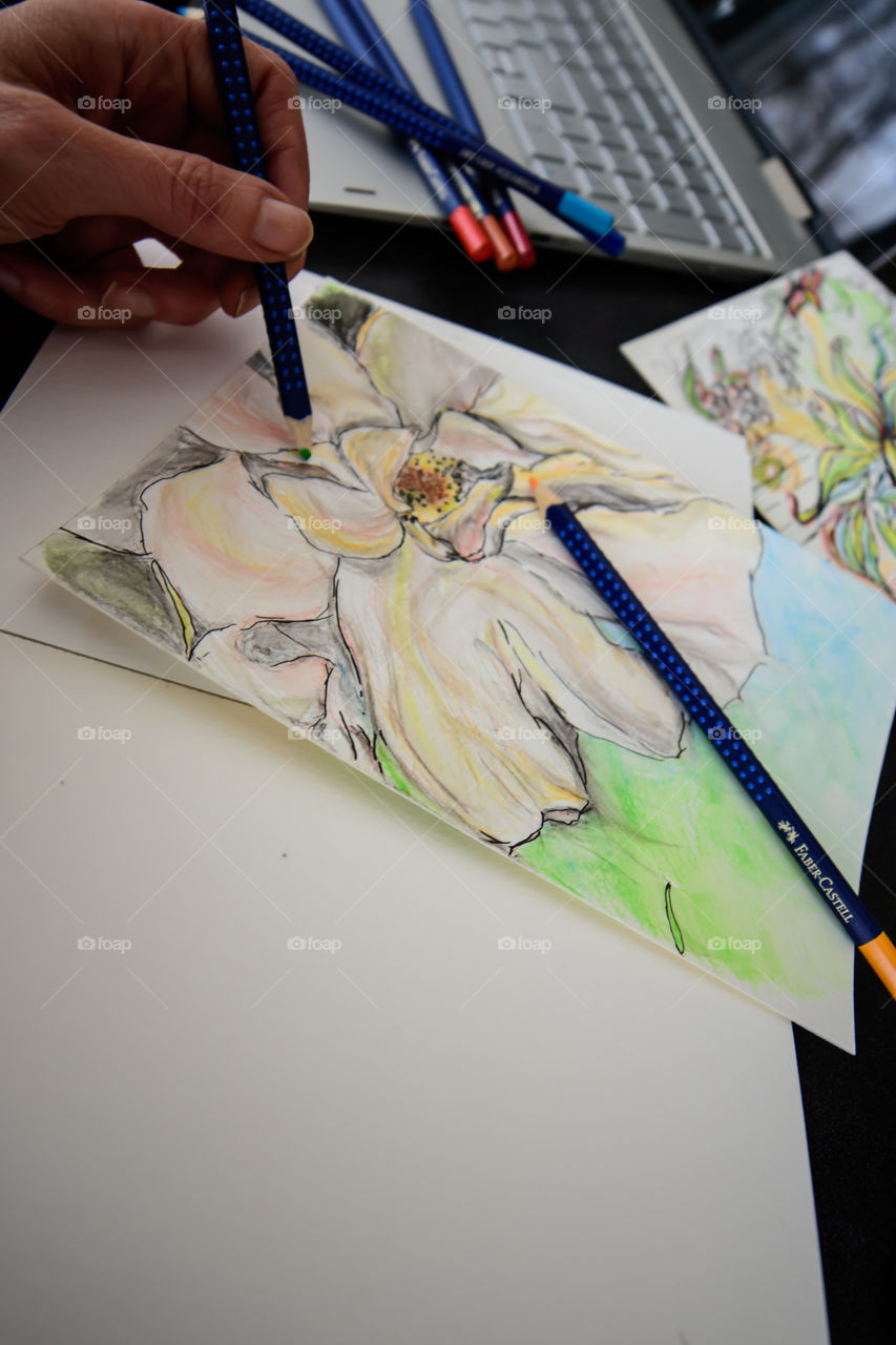 Low angle view of hand holding Faber-Castell Art Grip Aquarelle Colored watercolor pencils sketching closeup spring flower with sketchbook on table with laptop and coffee conceptual work life balance, creativity, lifestyle photography