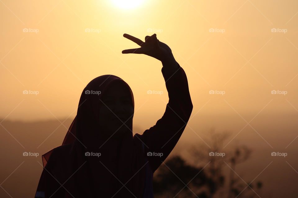 Silhouette of a girl with hijab showing peace hand sign with beautiful background of sunrise