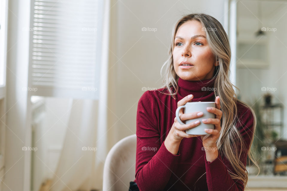 Young blonde woman girl with long hair in red sweater with cup of tea or coffee in hands in home