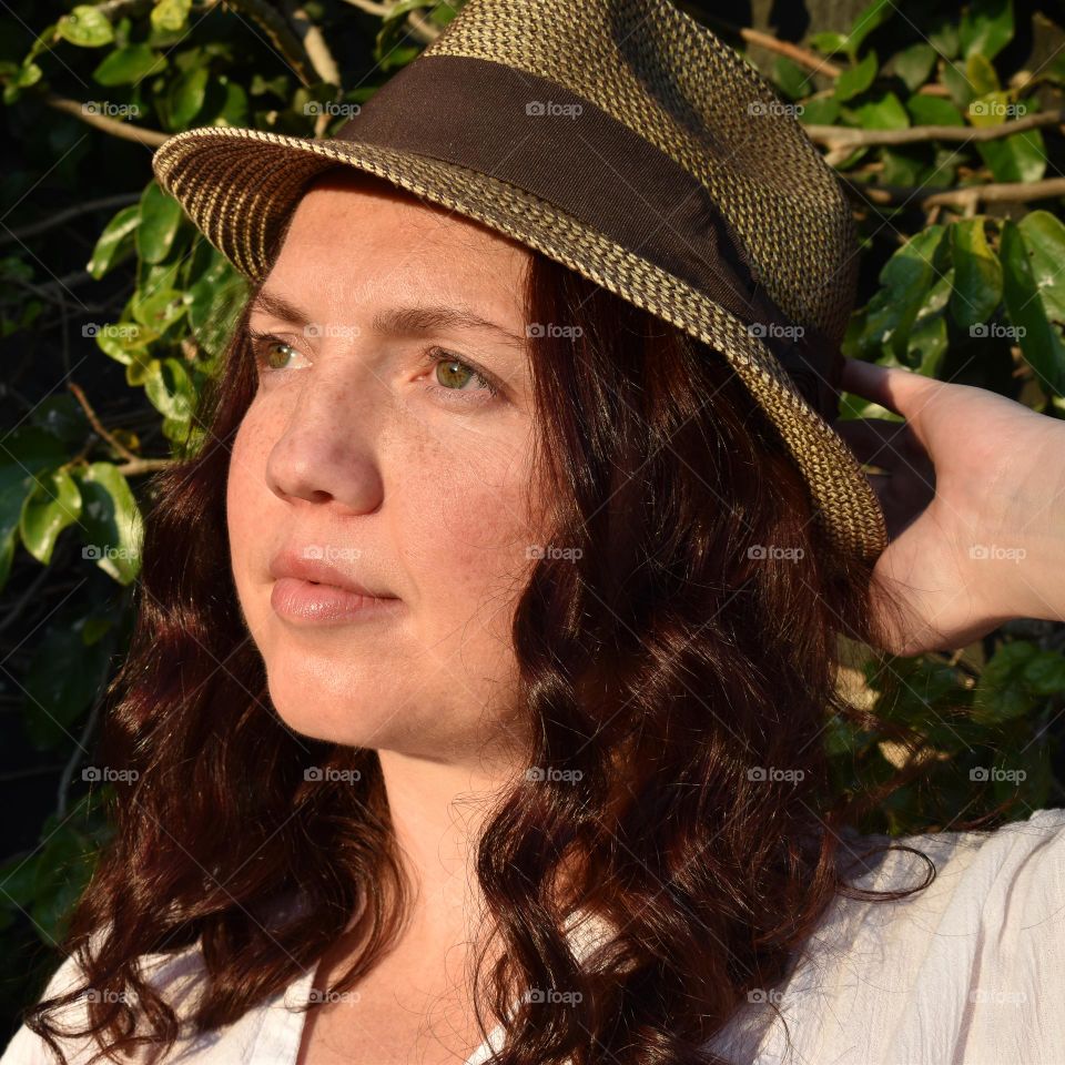 Women with natural and minimal makeup standing outdoors outside with wet hair. Women with red hair and green eyes standing next to tree taking a selfie. Sunlight shine on women hair and skin and making shadows. Women wearing a hat.