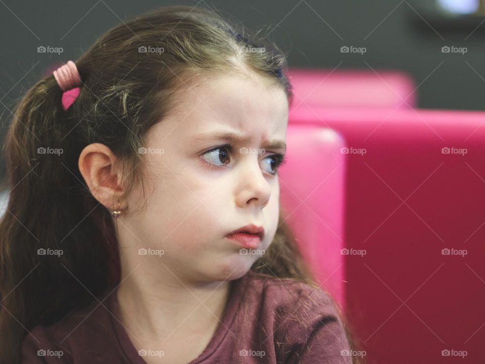 Portrait of one beautiful caucasian little brunette girl with ponytails on her head looks sadly and resentfully to the side, standing in a cafe in the afternoon, close-up side view. The concept of sad people and offended children.