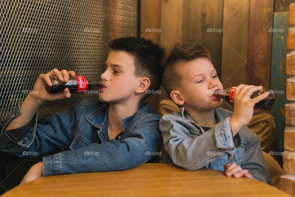 two lovely brothers, dressed in denim clothes, with beautiful hairstyles, are sitting in a cafe and drinking Coca-Cola.  communicate, laugh, smile.  two guys, two friends