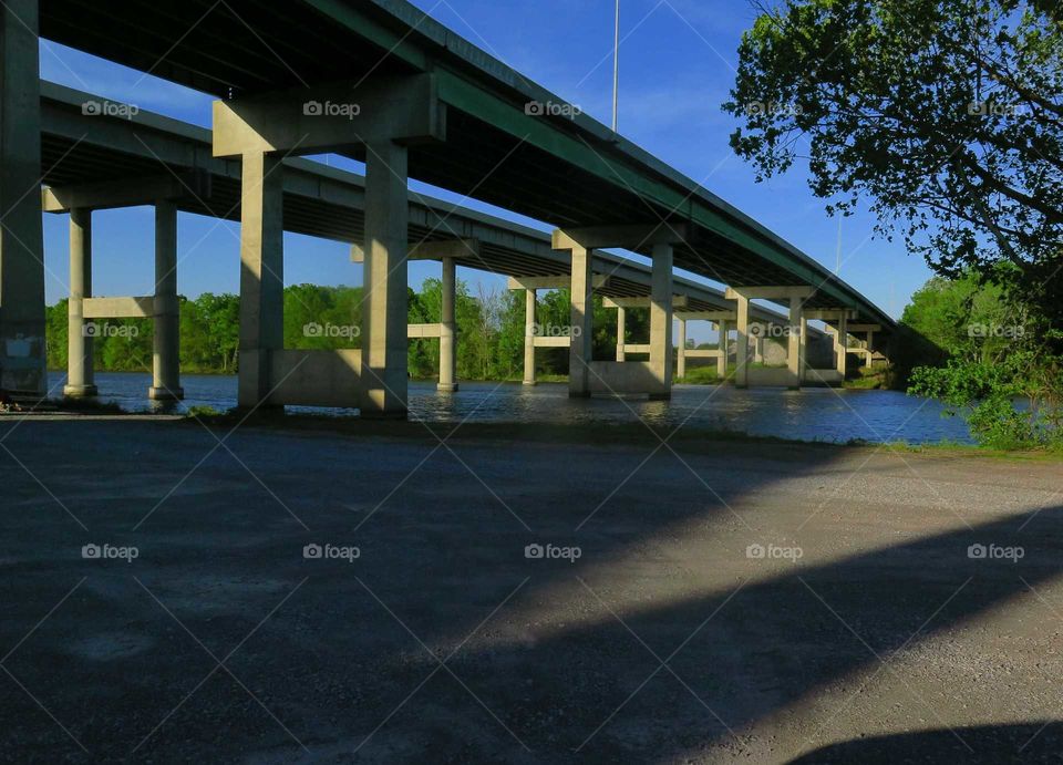 a bridge in Alabama casting Shadows on to the ground while crossing over a river the Coosa River to be exact as you see the Blue Sky in the background and a tree to the right cars pass over this bridge daily to and fro back and forth to work to home