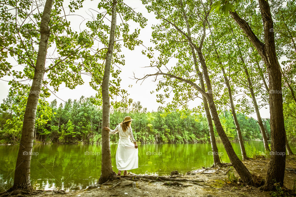 Woman from behind in white dress wearing straw hat, standing in front of the lake, in summer green forest landscape at afternoon, enjoying nature. Freedom, romance, resting and tranquility. Lonely female In thought, in the trees at the park.