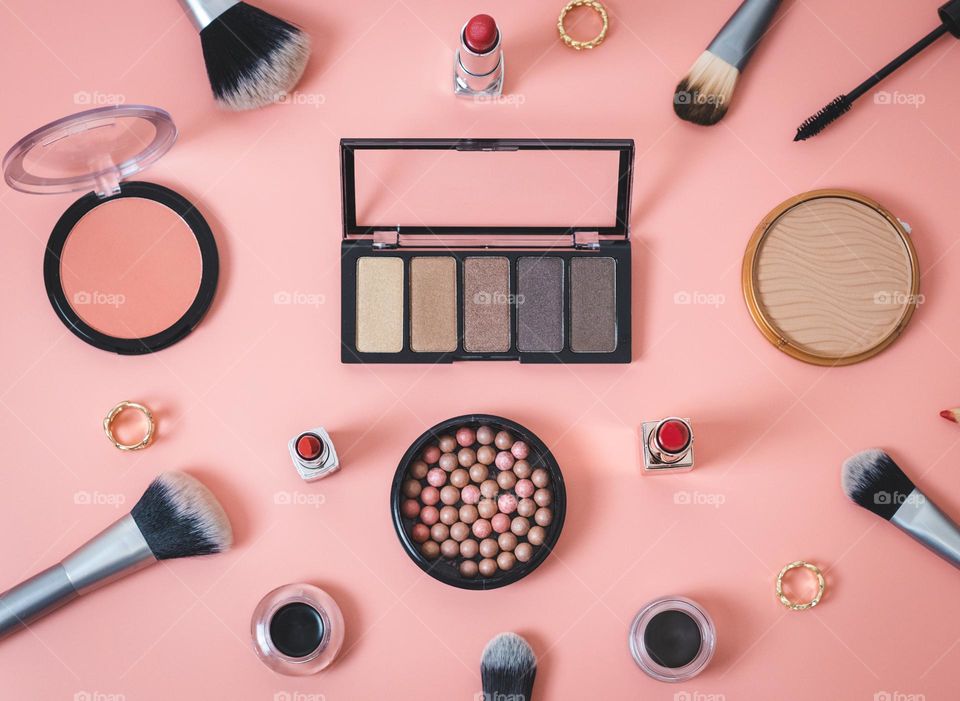 A set of cosmetics from an eyeshadow palette, nude face powders, red lipsticks, soft dark eyebrow shadows, brushes, brushes, pencils and rings lie scattered on a pink background, lie flat on a close-up. The concept of female cosmetics, beauty salon.