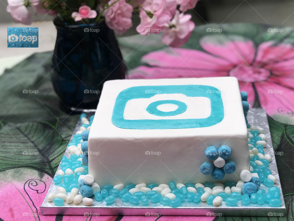 A foap wedding cake with the logo made of white & turquoise fondant. Daisies of turquoise gummies & marshmallows on the side of the cake & turquoise & white jelly beans on the cake base.  All on a green & pink table cloth with a vase of pink roses. 😋