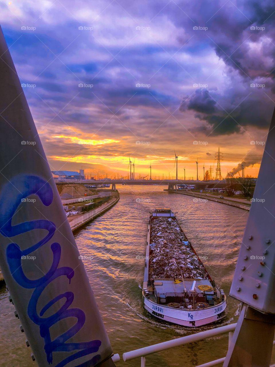 Large river transport boat going underneath a bridge over the river scheldt at sunset showing the Golden glow in the sky under the dark and blue sky above