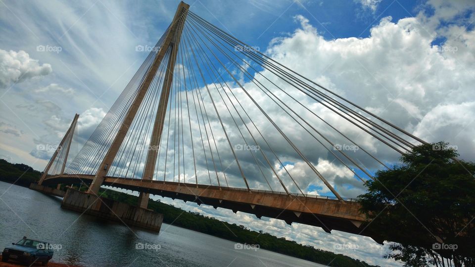 O céu nublado e a ponte sobre um rio, suspensa, de concreto e cabos. Divisa de Estados. MS e MG, Brasil.