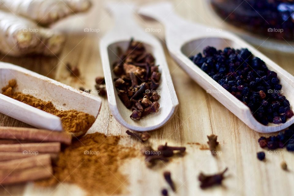 Closeup of ground cinnamon and cinnamon sticks, ginger root, whole cloves, and dried elderberries in wooden scoops on a wooden surface as ingredients for homemade elderberry syrup