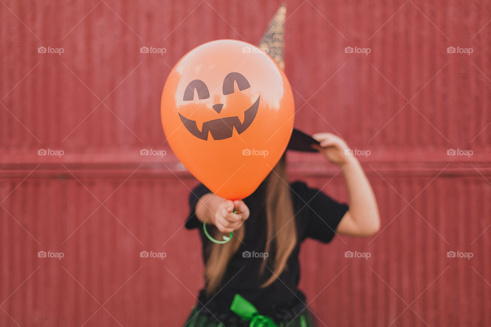 A little girl dressed as a witch for halloween holds a pumpkin-shaped balloon in her hands.